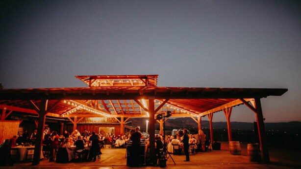 Un área de comedor al aire libre cubierta, con estructura de madera y mesas, frente a un cielo nocturno con una luna visible.
