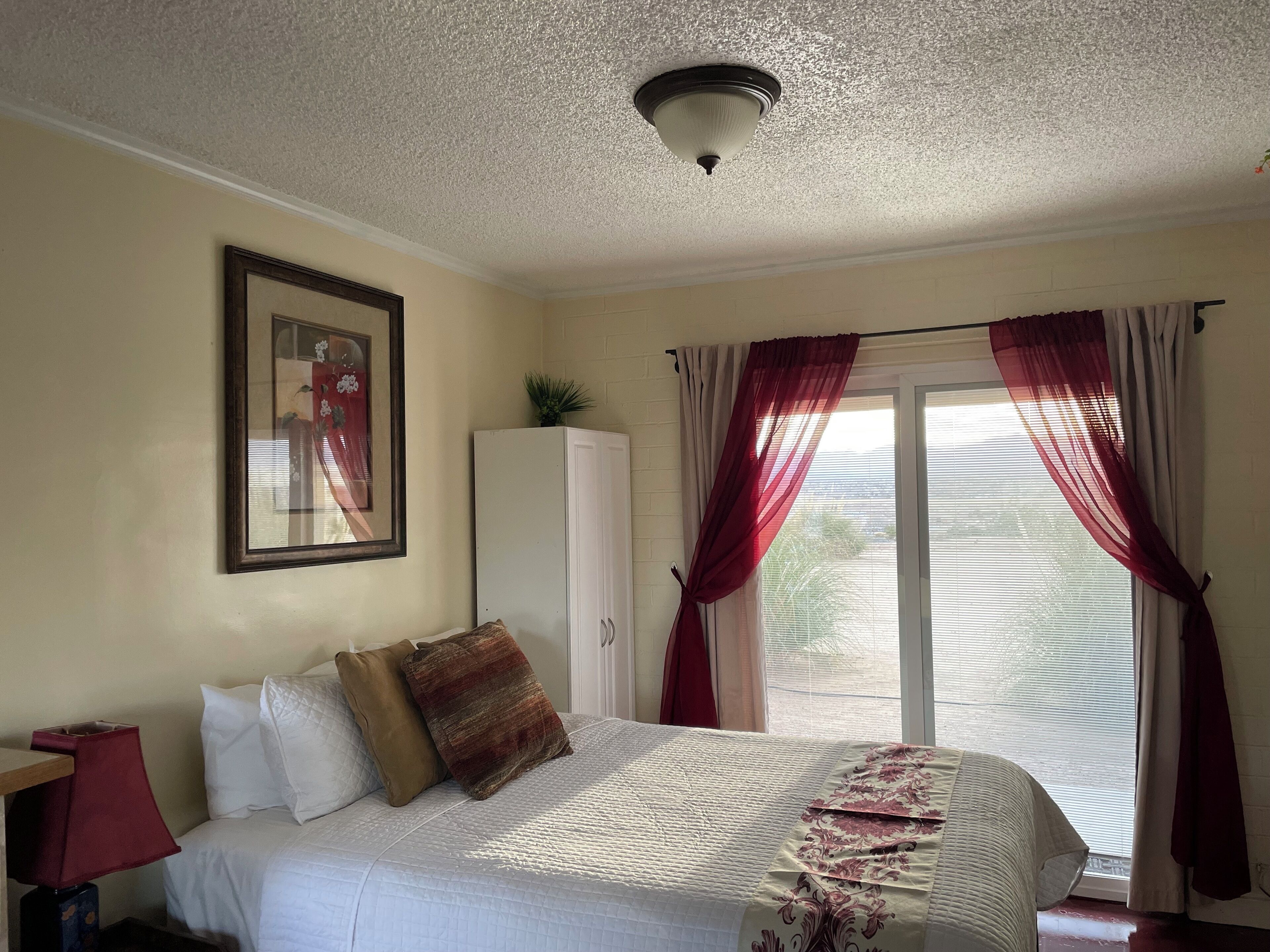 Un dormitorio con cama, armario, un cuadro en la pared y ventana con cortinas.