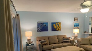 A living room with a sofa, ottoman, and a small table with a lamp. There are two wall art pieces and a ceiling fan. - Beach Lovers, Oceanview, Cute 1 BR, 2 full BA condo in Surf City sleeps 5 (Surf City)