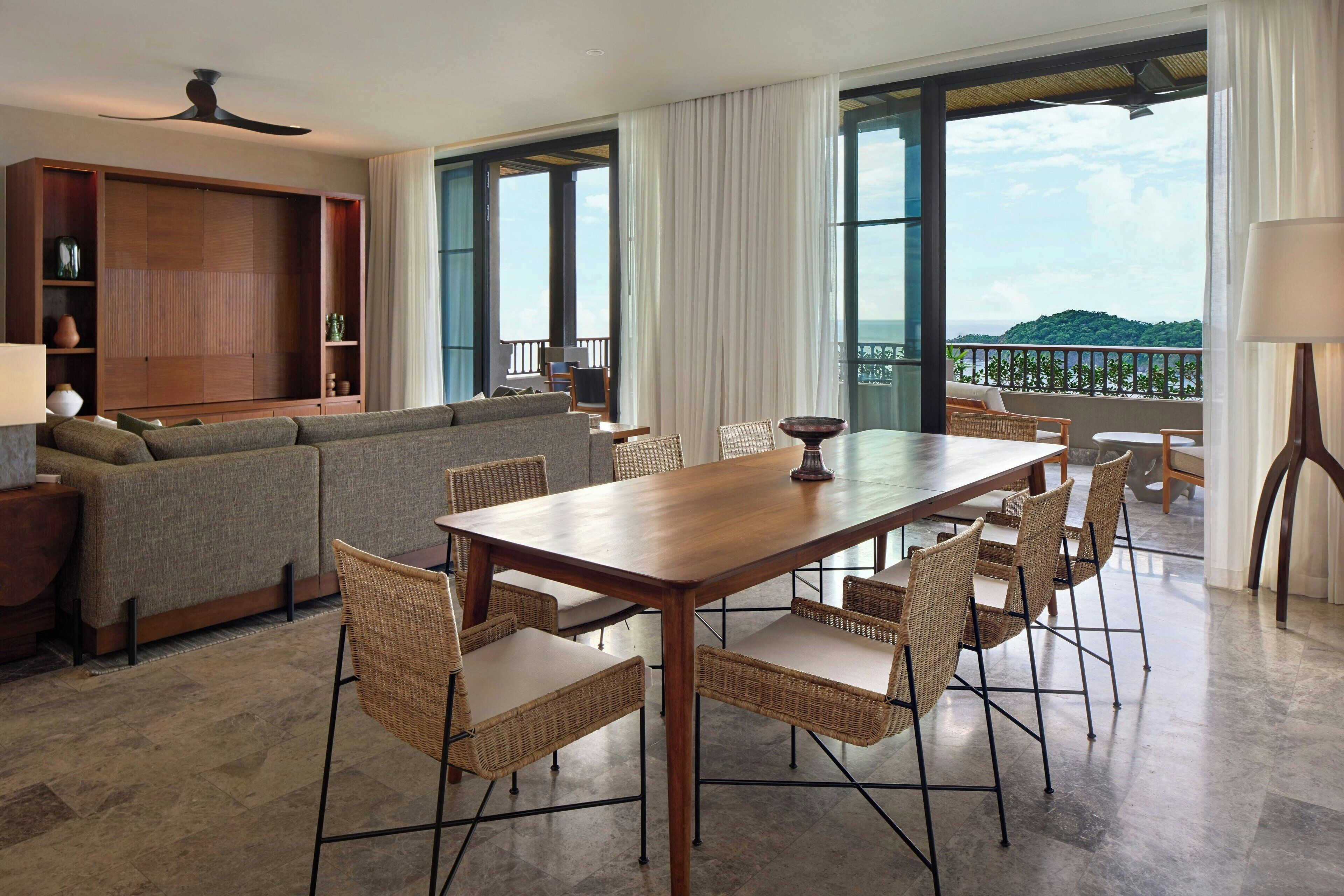 Un amplio salón con una mesa de comedor de madera, sillas de mimbre, un sofá gris y un gran ventanal con vistas a las colinas.