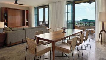Un amplio salón con una mesa de comedor de madera, sillas de mimbre, un sofá gris y un gran ventanal con vistas a las colinas.