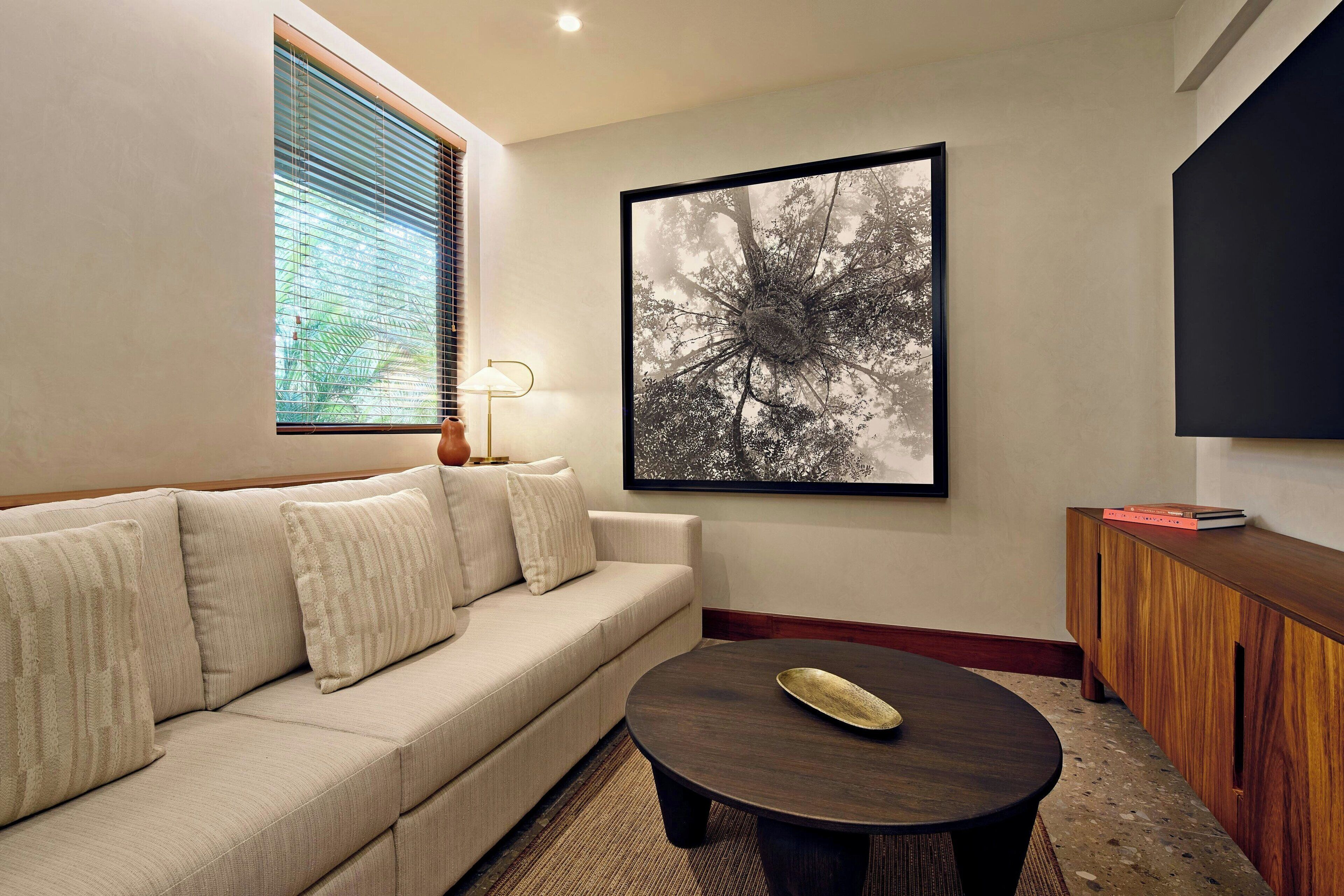 A modern living room with a beige sofa, a round coffee table, a framed black and white tree photograph on the wall, a floor lamp, and a wooden sideboard with books.