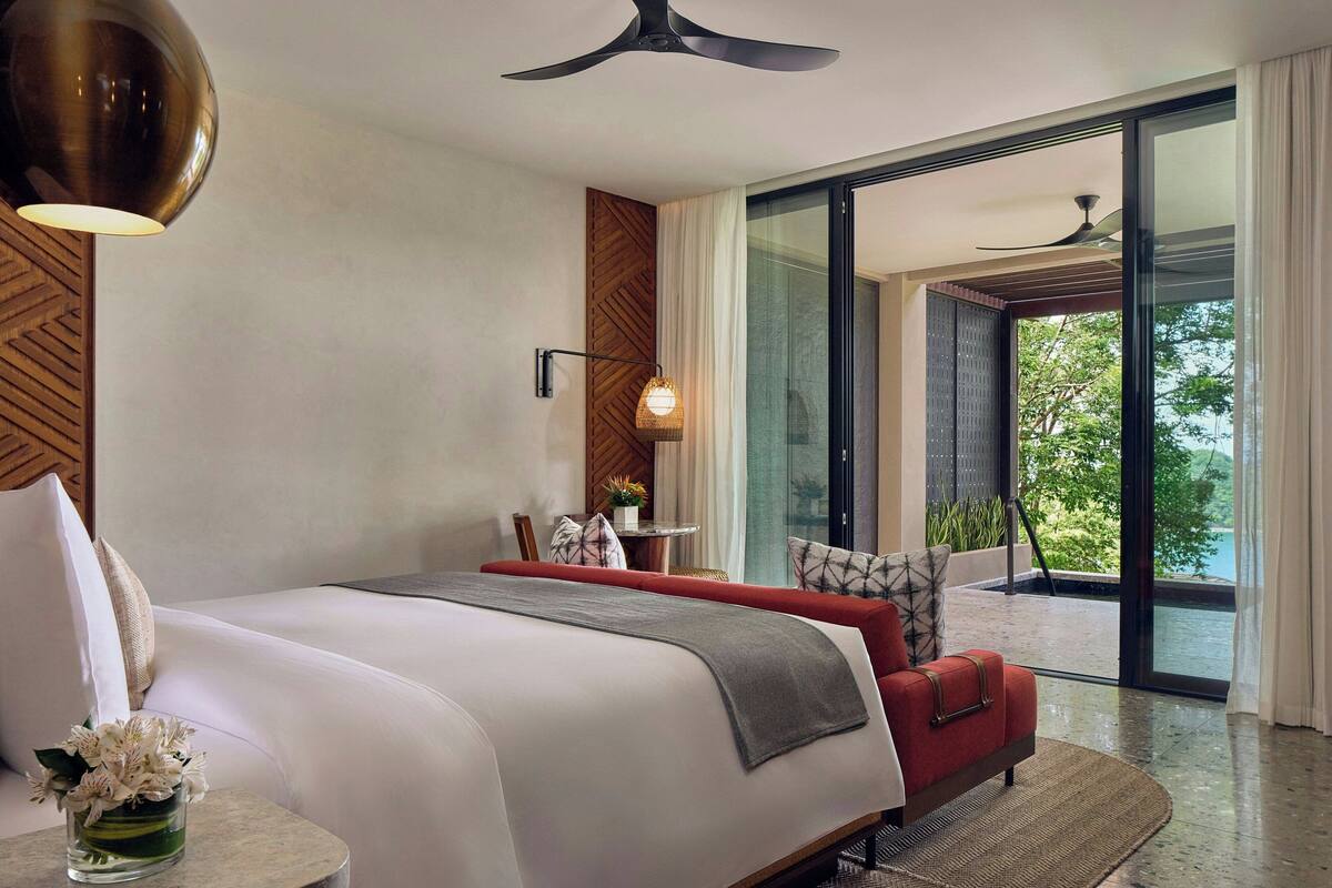 A modern hotel room with a large bed, a red sofa, a dining table with chairs, and a view of the outdoors through a sliding glass door.