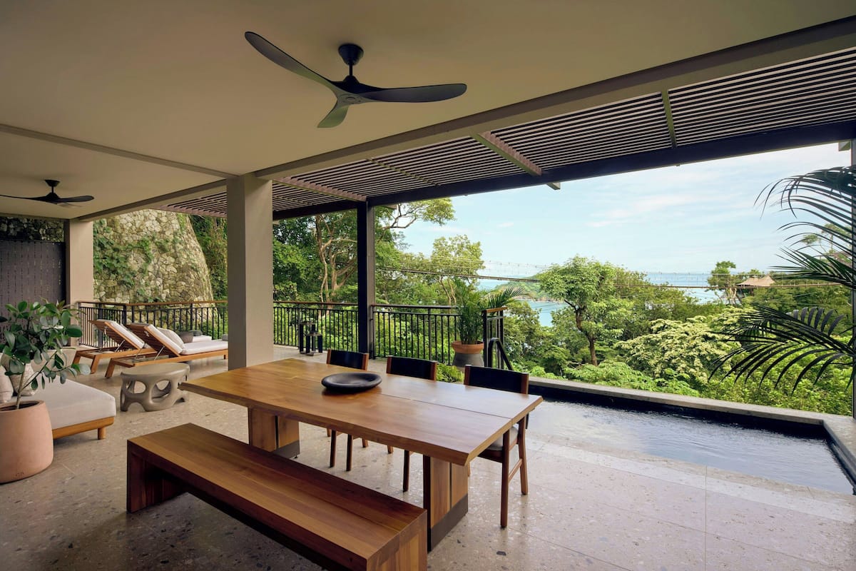 A spacious outdoor area with a wooden table and benches, surrounded by potted plants and a view of lush greenery and a body of water.