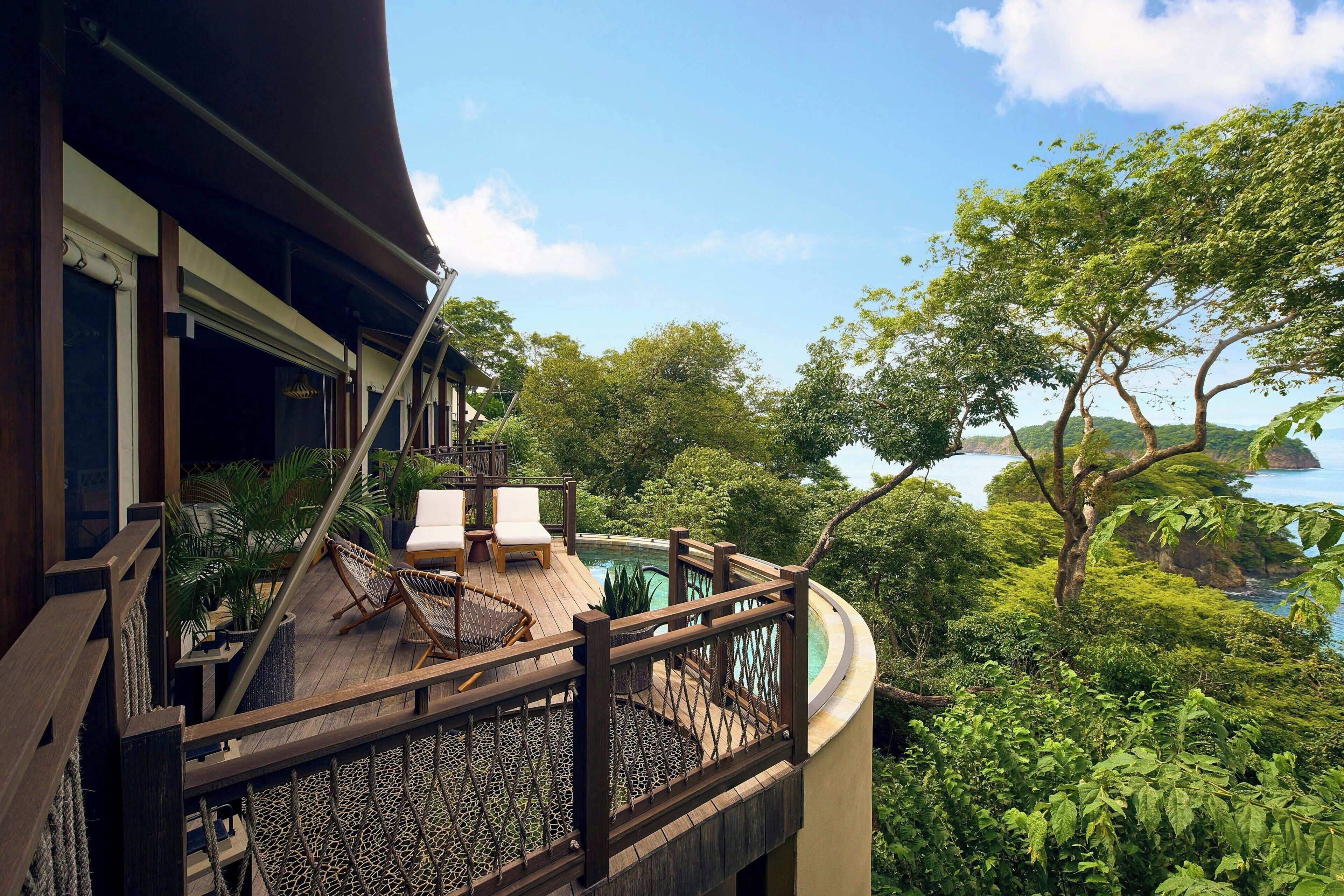 A wooden deck with a pool, surrounded by lush greenery and overlooking a coastal area.