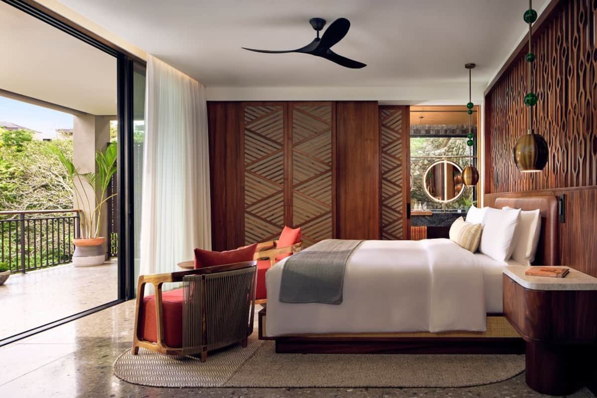 A bedroom with a bed, a chair, a bedside table, a ceiling fan, and a view of the outdoors.