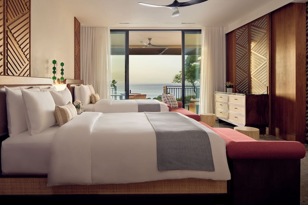 A hotel room with two beds, a balcony, a ceiling fan, and a view of the ocean.
