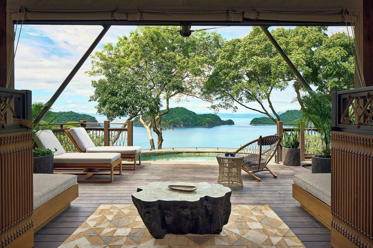 A deck with lounge chairs, a coffee table, and a view of a body of water and greenery.
