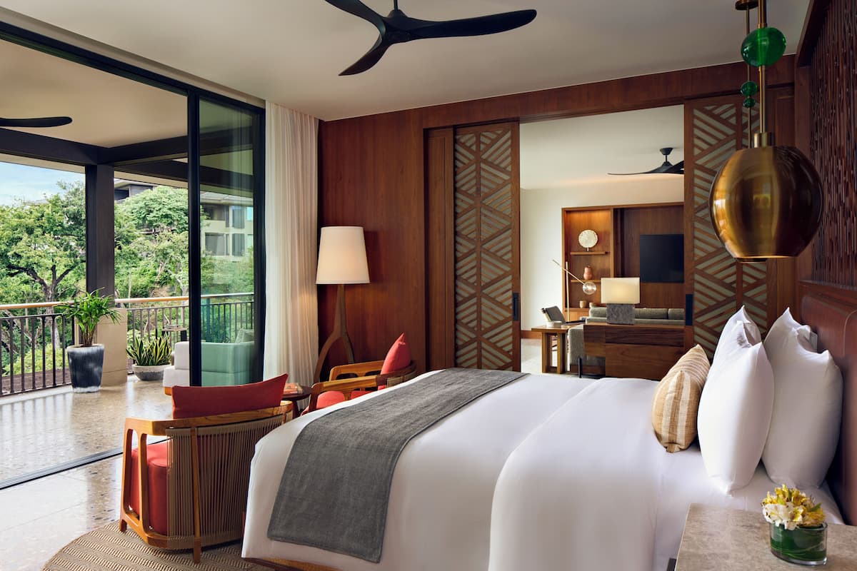 A hotel room with a large bed, a desk, a chair, a balcony with plants, and a view of the outdoors.