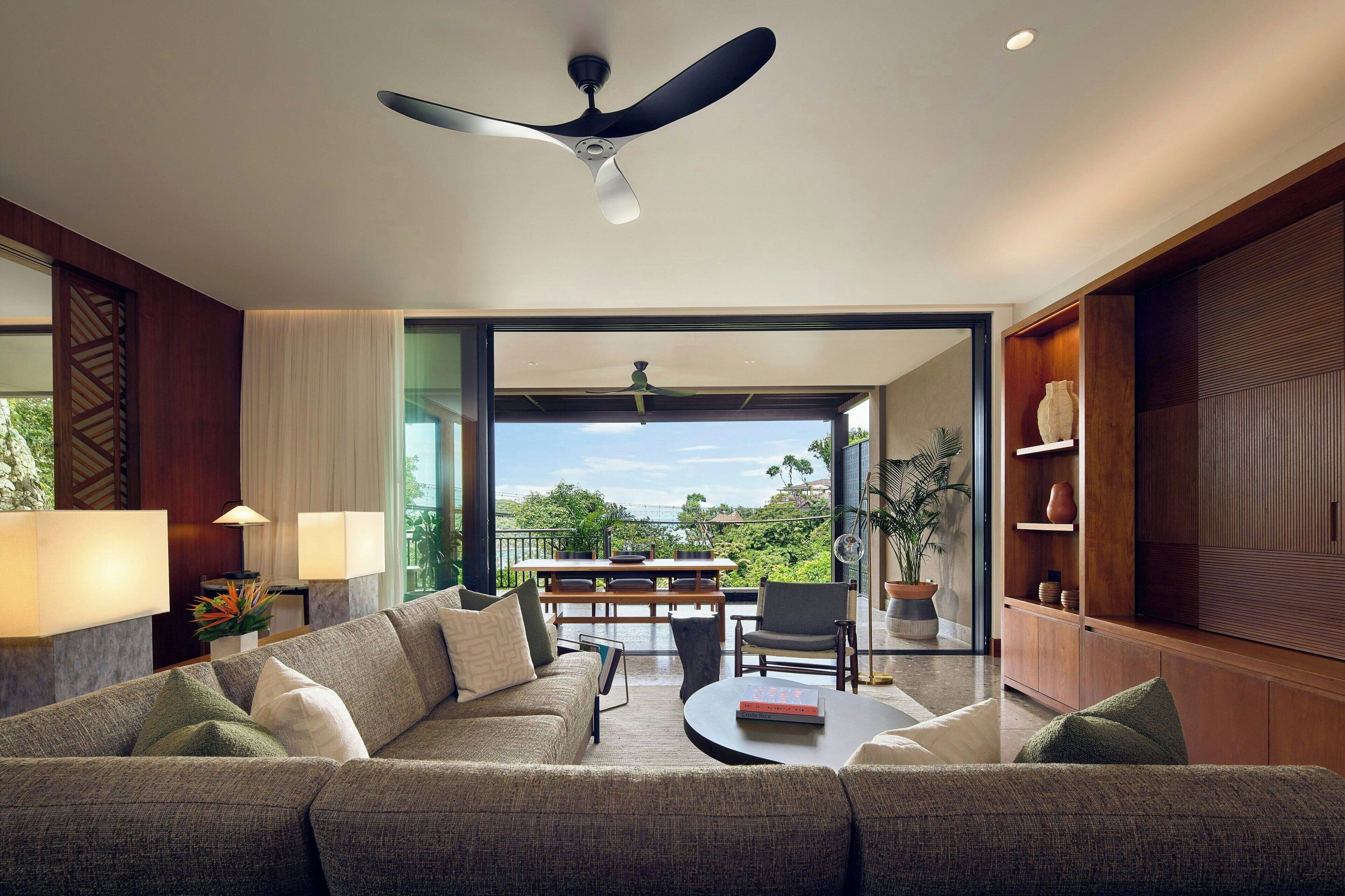Una sala de estar moderna con un sofá, una mesa de centro y un comedor con una mesa y sillas. Hay un ventilador de techo, un ventanal con vistas al exterior y una estantería con objetos decorativos.