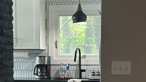 A modern kitchen with white cabinets, a black pendant light, a window with a view of trees, and a small wooden bowl on a stand.