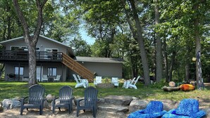 A house with a deck and a staircase leading to a second floor. There are several Adirondack chairs and a blue inflatable lounge chair on the lawn.