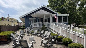 Un espace extérieur aménagé avec une zone de repos couverte, comprenant des fauteuils Adirondack et une petite table, un escalier menant à un porche et un drapeau américain.