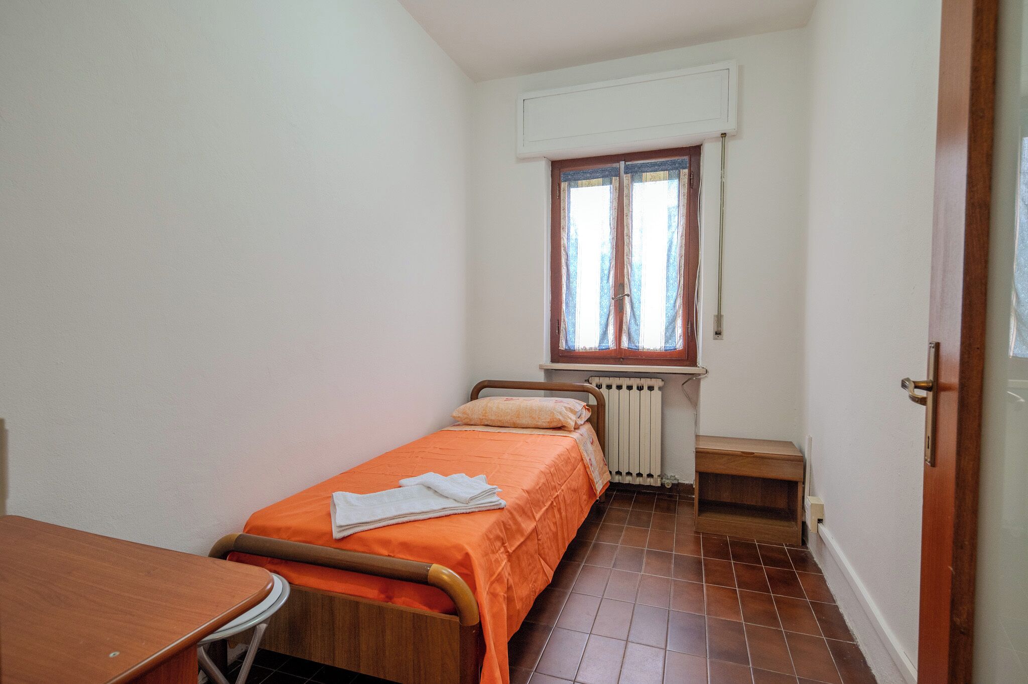 Une petite chambre avec un lit simple, un lit en bois, une couverture orange, un bureau en bois et une fenêtre avec un store blanc.