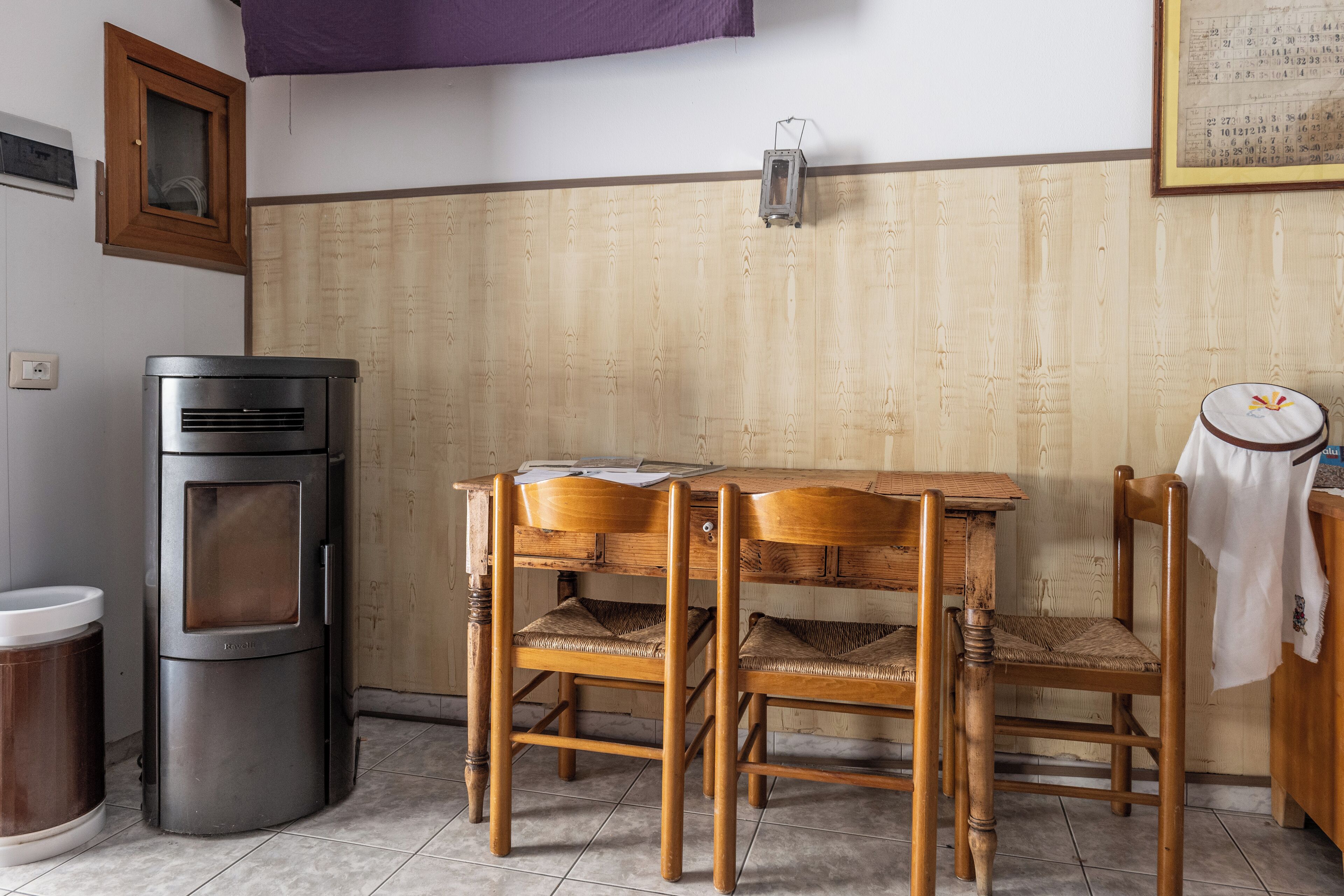 Una cocina con estufa, una mesa, dos sillas, un bote de basura y una lámpara fijada a la pared.