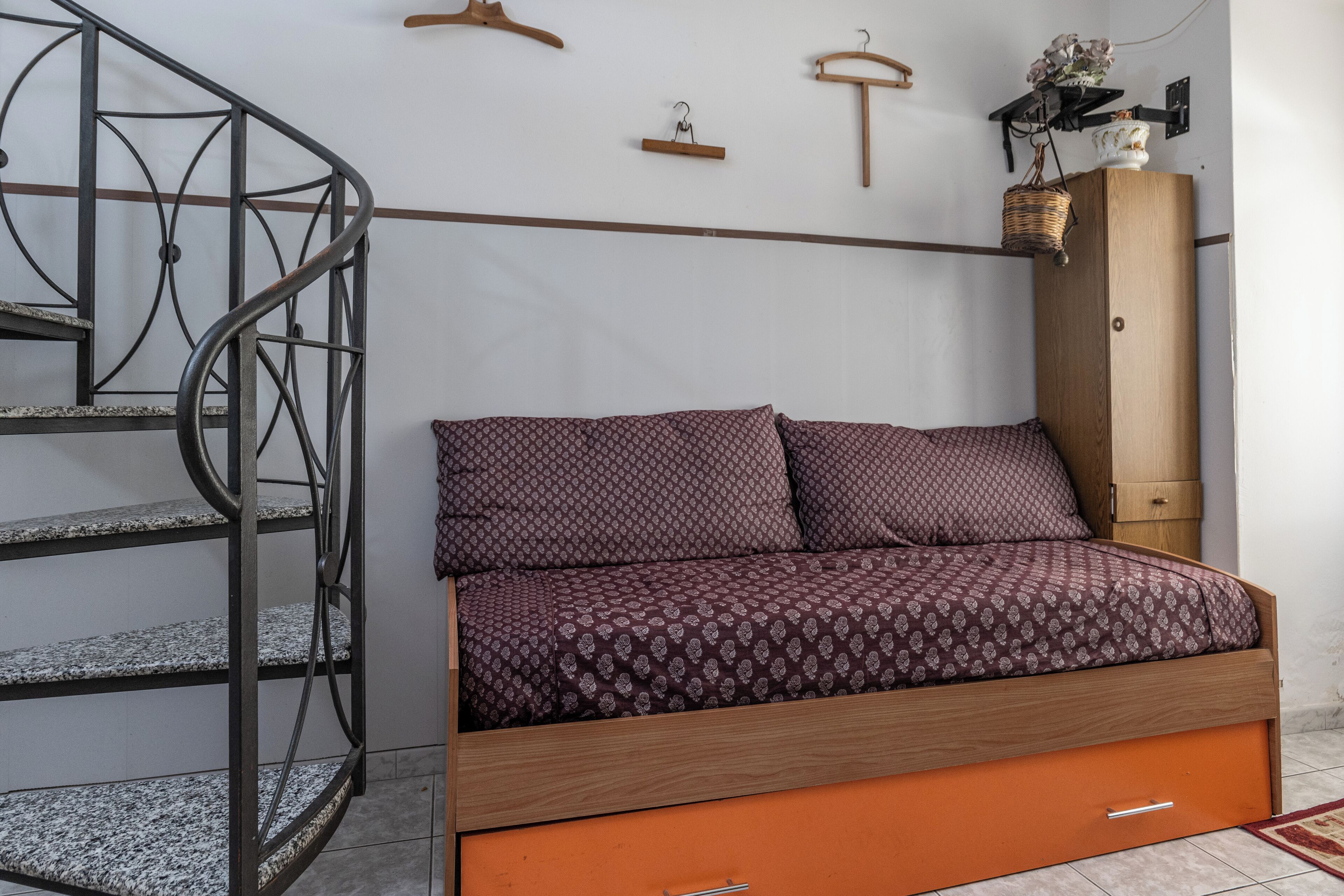 A bedroom with a bed, a wooden wardrobe, a metal spiral staircase, and a wall-mounted shelf.