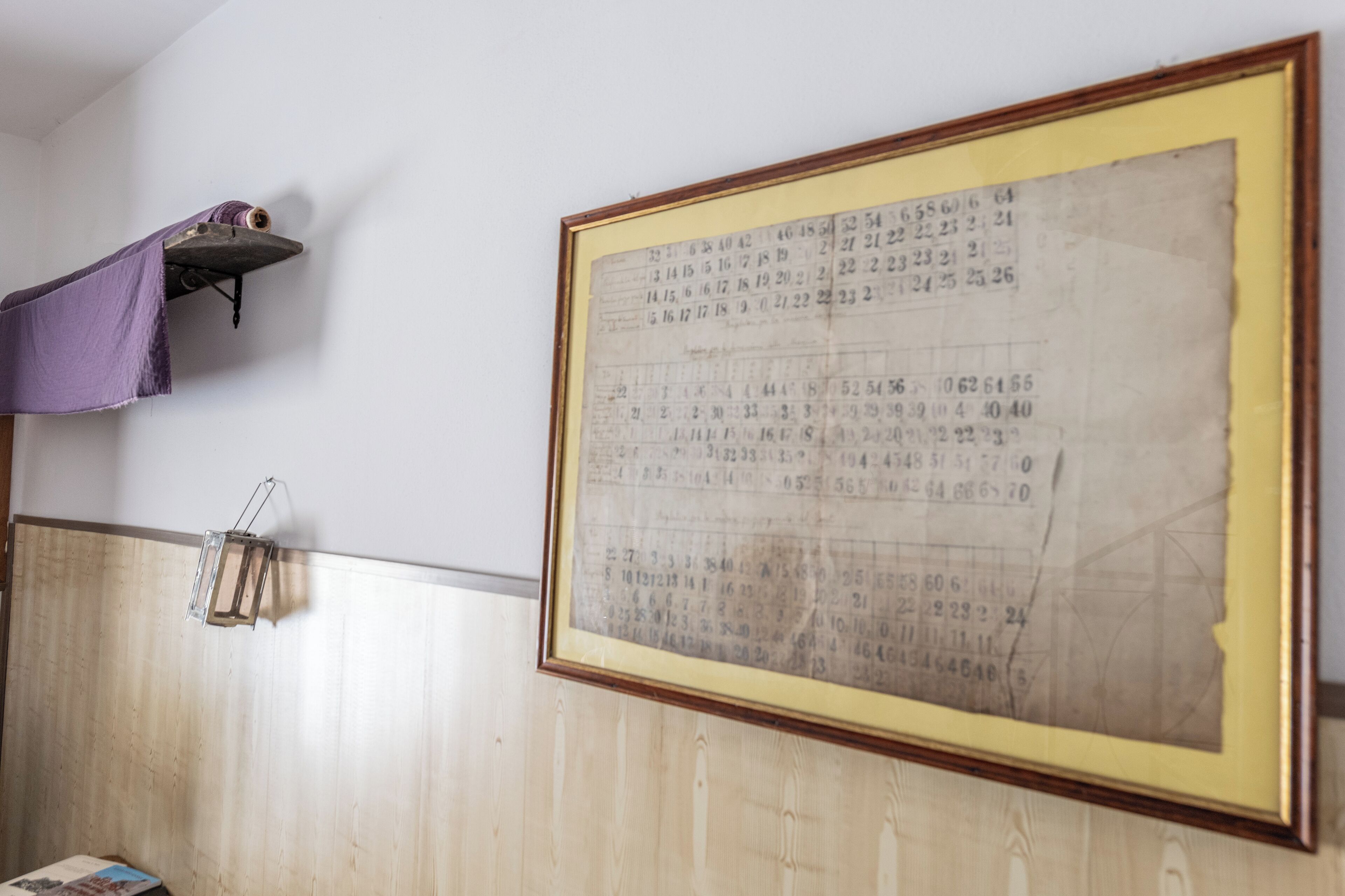 A room with a large framed document on the wall, a wooden panel wall, a shelf with a purple cloth, and a small table with a book.