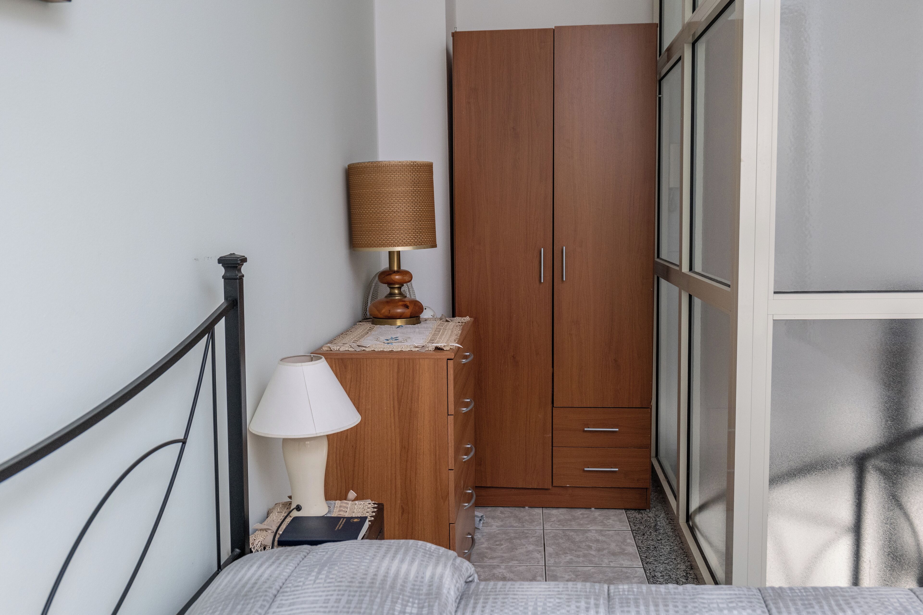 A bedroom with a bed, bedside table, lamp, wardrobe, and a window with blinds.