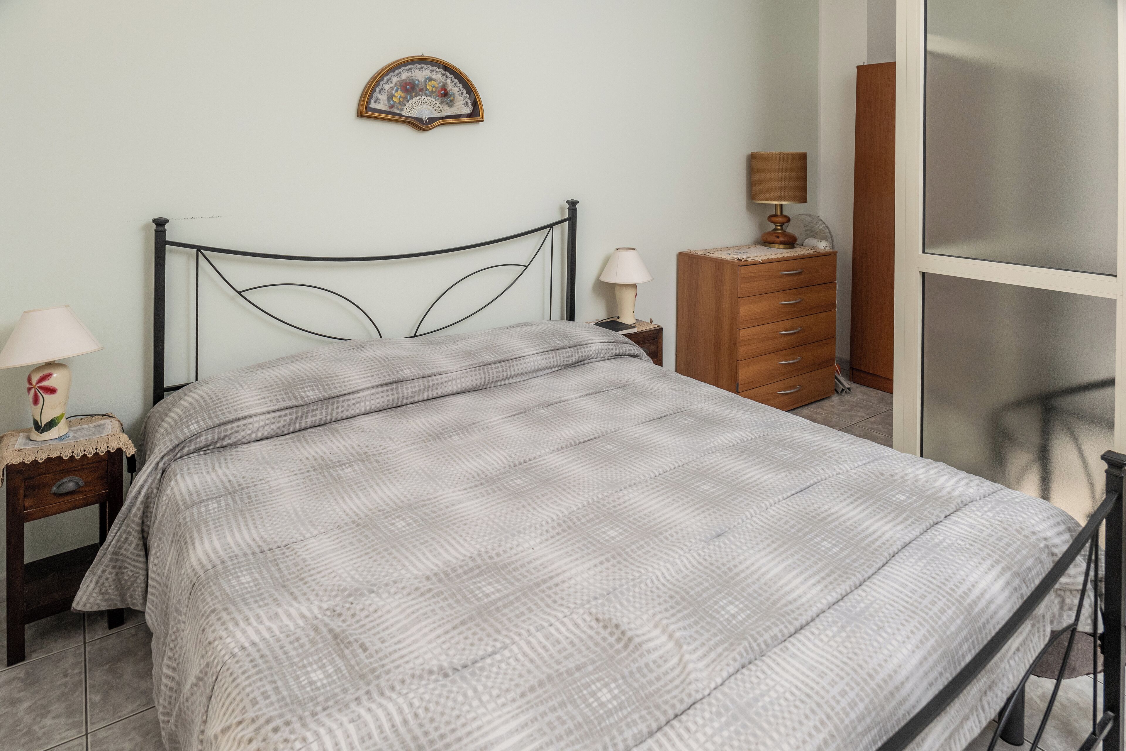 A neatly made bed with a checkered bedspread, a bedside table with a lamp, a wooden dresser, and a wall-mounted decorative item.