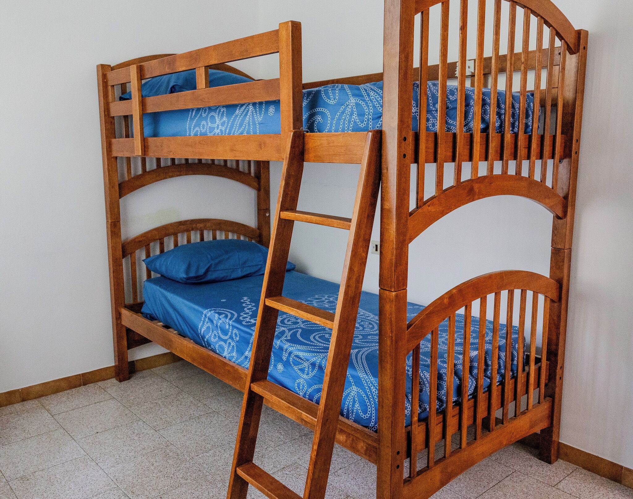 Una cama litera con escalera de madera, ropa de cama azul estampada y una pared blanca al fondo.