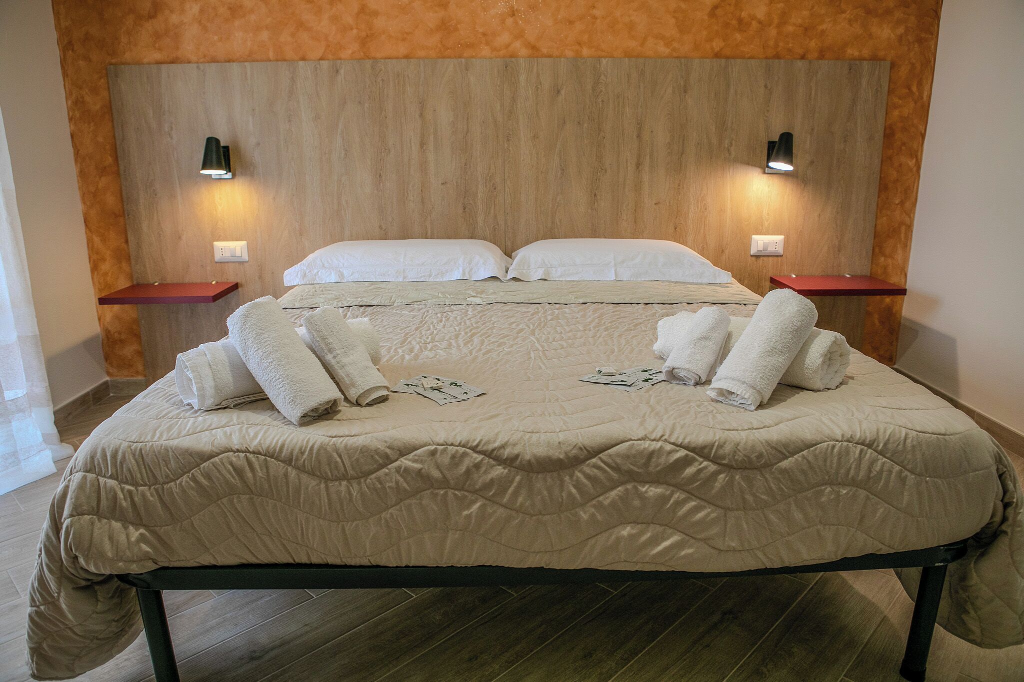 Un llit ben fet amb un llençol beix, dos coixins blancs i diverses tovalloles doblegades amb cura. Hi ha dues taules de nit amb superfície vermella i làmpades negres. La habitació té un paviment de fusta i un capçal de llit de fusta.