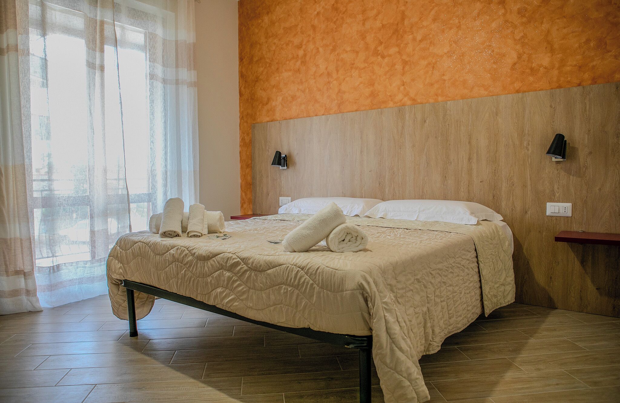 Un letto rifatto con una coperta beige, due cuscini e asciugamani arrotolati su un pavimento in legno.