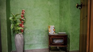 A corner with a green textured wall, a tall vase with flowers, and a small wooden side table with a picture frame and a book.