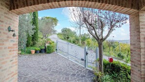 A stone-paved courtyard with a metal gate, surrounded by greenery and a view of a road and trees. - A Casa Di Lea (Mondavio)