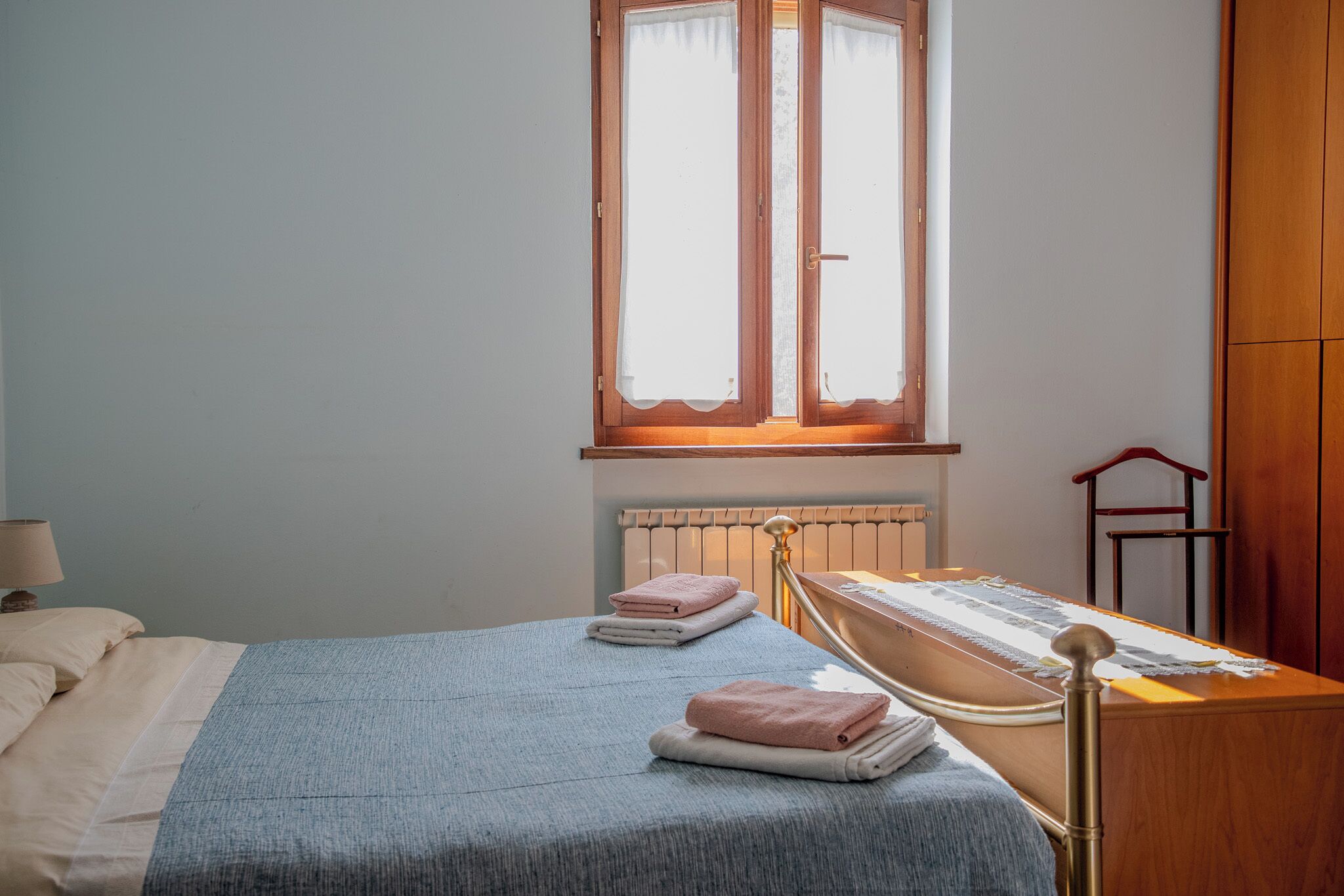 A neatly made bed with a blue blanket and two folded towels, a wooden bedside table with a lamp, and a window with curtains.