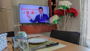 Una televisione che mostra un quiz, un tavolo da pranzo con un piatto e posate, e un vaso con fiori artificiali.