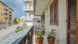 Un balcone con piante in vaso, vista sulla strada e un edificio con balconi e persiane.