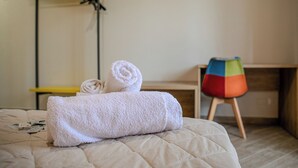 Un letto rifatto con cura, asciugamani bianchi arrotolati, una scrivania in legno e una sedia colorata in una stanza dal design essenziale e pulito.