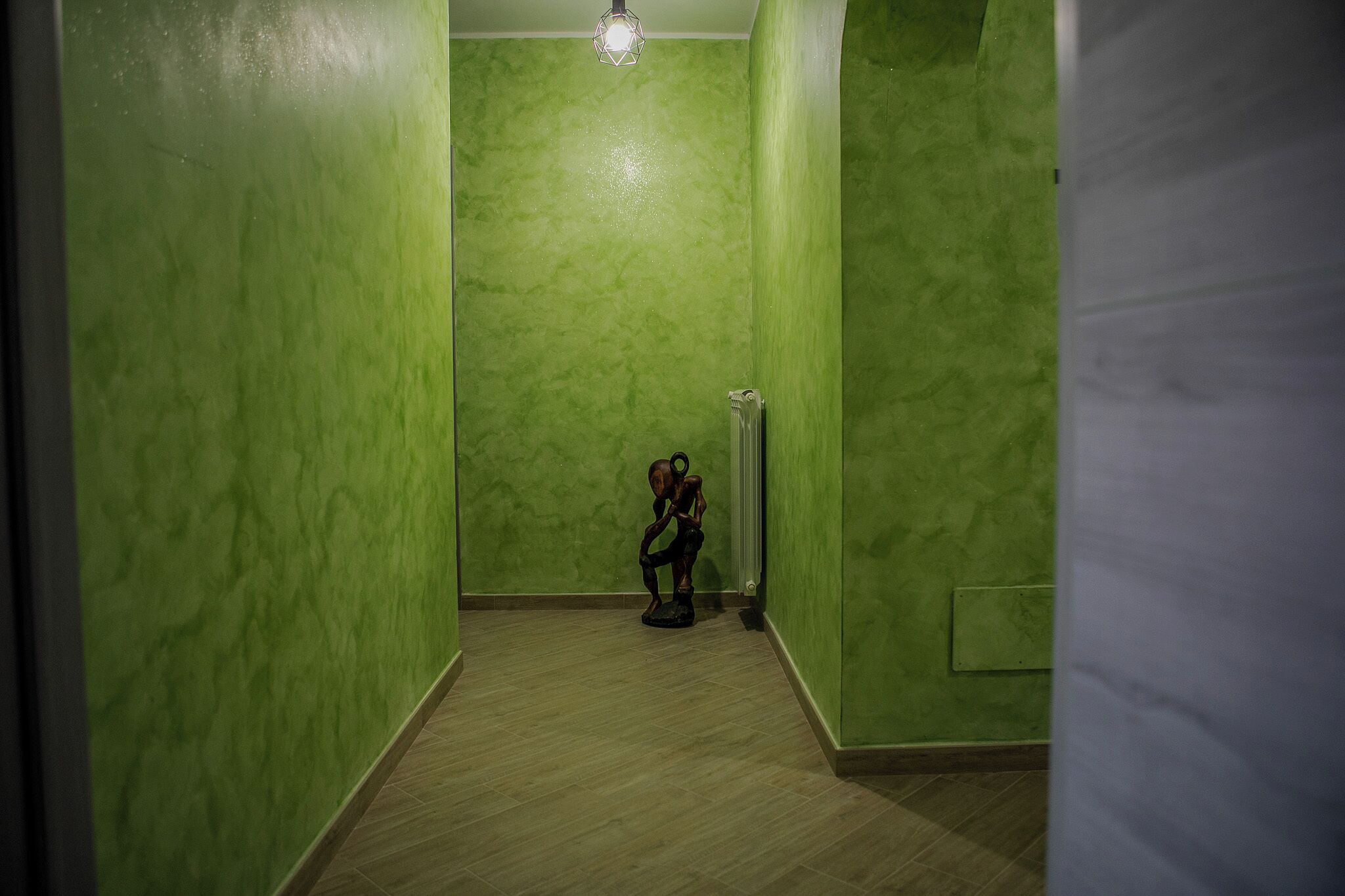 Un pasillo estrecho con paredes verdes y suelo de madera. Un radiador está fijado a la pared, y una persona sentada en una silla en el centro del pasillo.