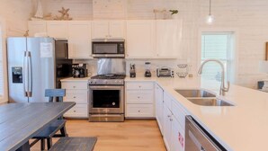 A modern kitchen with white cabinetry, stainless steel appliances, and a wooden dining table. - Driftwood Delight *private Pool!*Beachway Bungalow (Port Aransas)