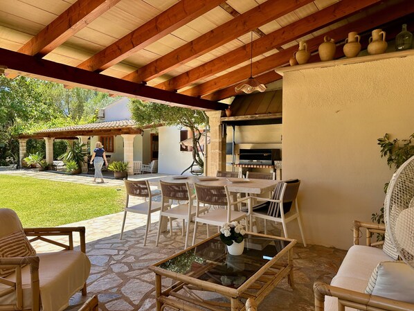An outdoor patio with stone flooring, white wicker furniture, a glass-top table with flowers, and a stone oven.