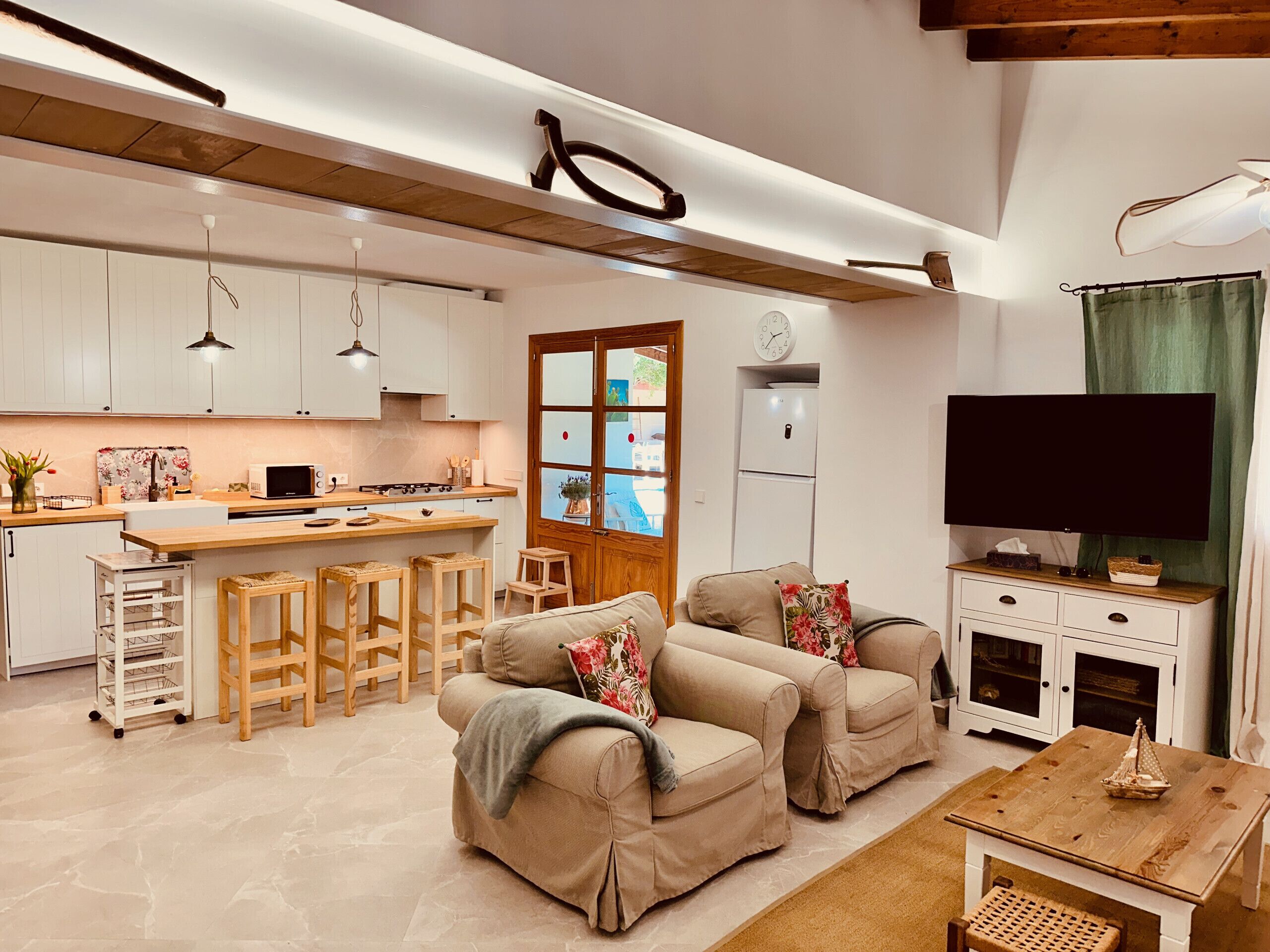 A modern kitchen with white cabinets, a central island, and bar stools. There is a living area with two beige sofas and a television.