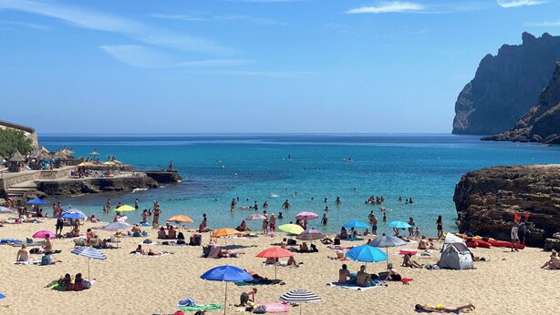 Une plage bondée où les gens se prélassent au soleil et se baignent, des tentes sont installées, et une falaise rocheuse se dresse à l'arrière-plan.