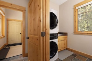 A laundry room with a front-loading washer and dryer, wooden cabinets, a window with a view of trees, and a door leading to another room.