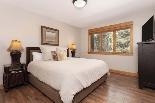 Une chambre à coucher avec un grand lit, deux lampes de chevet, un tableau encadré au mur, une fenêtre donnant sur les arbres et une commode en bois foncé.