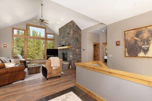 A spacious living area with a stone fireplace, large windows, and a wooden staircase. - Lovely renovated home w/ hot tub & stunning views (Big Sky)