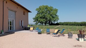 A building with a beige exterior, a large tree in the background, and several outdoor chairs and tables. - Champ Clair - SAINT BENIGNE (SAINT BENIGNE)