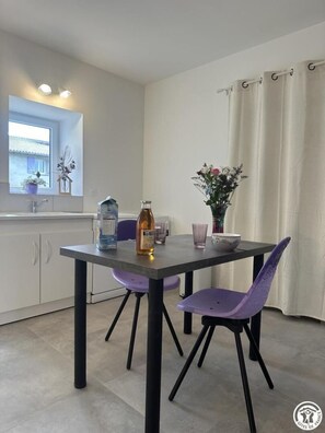 Une cuisine moderne avec une table à manger, des chaises violettes, une bouteille d’huile d’olive et un vase rempli de fleurs.
