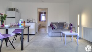 Ein modernes Wohnzimmer mit grauem Sofa, einem weißen Couchtisch und violetten Stühlen. Im Hintergrund ist ein Küchenbereich mit Kühlschrank, Mikrowelle und einem Esstisch mit violetten Stühlen erkennbar.