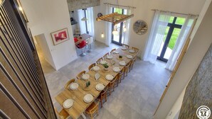 Un coin repas avec une longue table en bois dressée pour le repas, une cuisine en arrière-plan et de grandes fenêtres avec des rideaux.