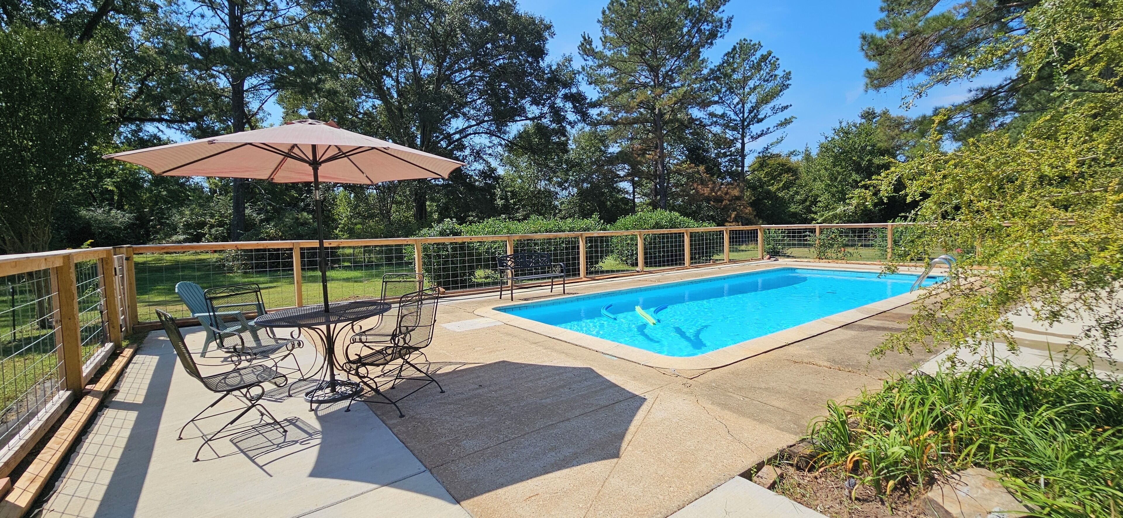 A rectangular swimming pool with a wooden deck, surrounded by a metal fence. There is a table and chairs with a metal umbrella nearby.