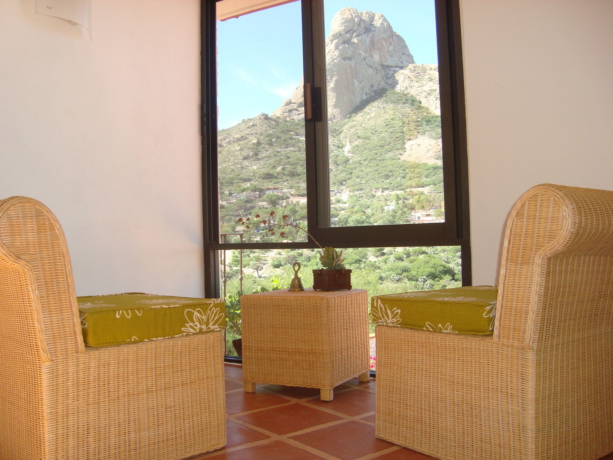 A room with two wicker chairs and a small table with a plant, overlooking a mountainous landscape through a large window.