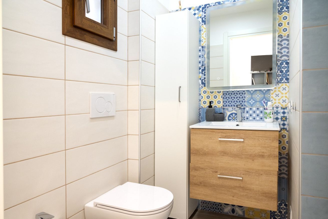 A bathroom with a white toilet, a wooden cabinet, and a tiled wall with a blue and yellow pattern.