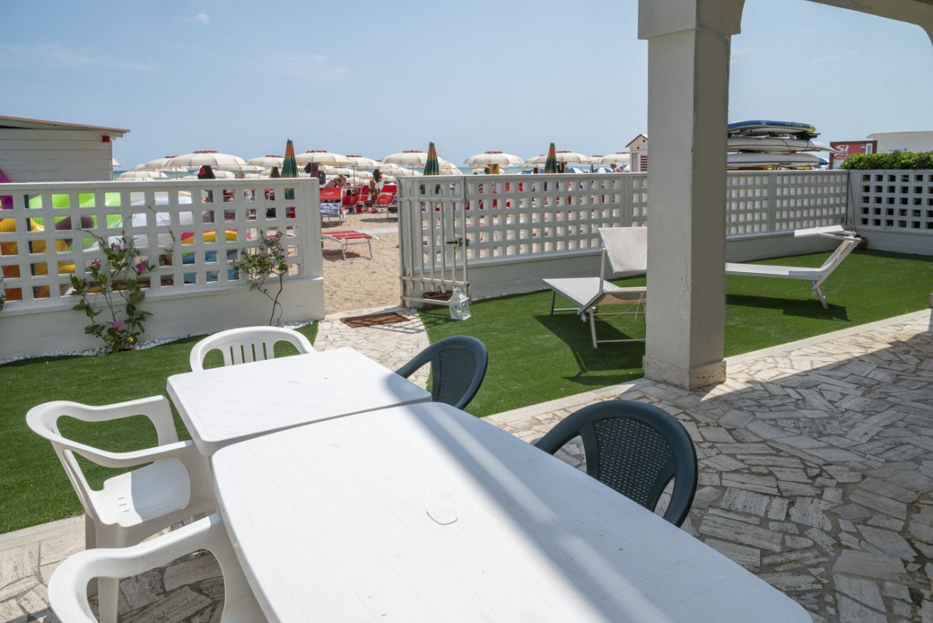 Un espace extérieur aménagé en terrasse, avec une table et des chaises blanches, une clôture blanche et, en arrière-plan, une vue sur des parasols de plage.
