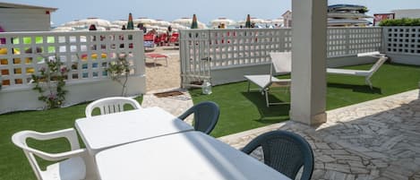 An outdoor patio area with a white table and chairs, a white fence, and a view of beach umbrellas in the background.