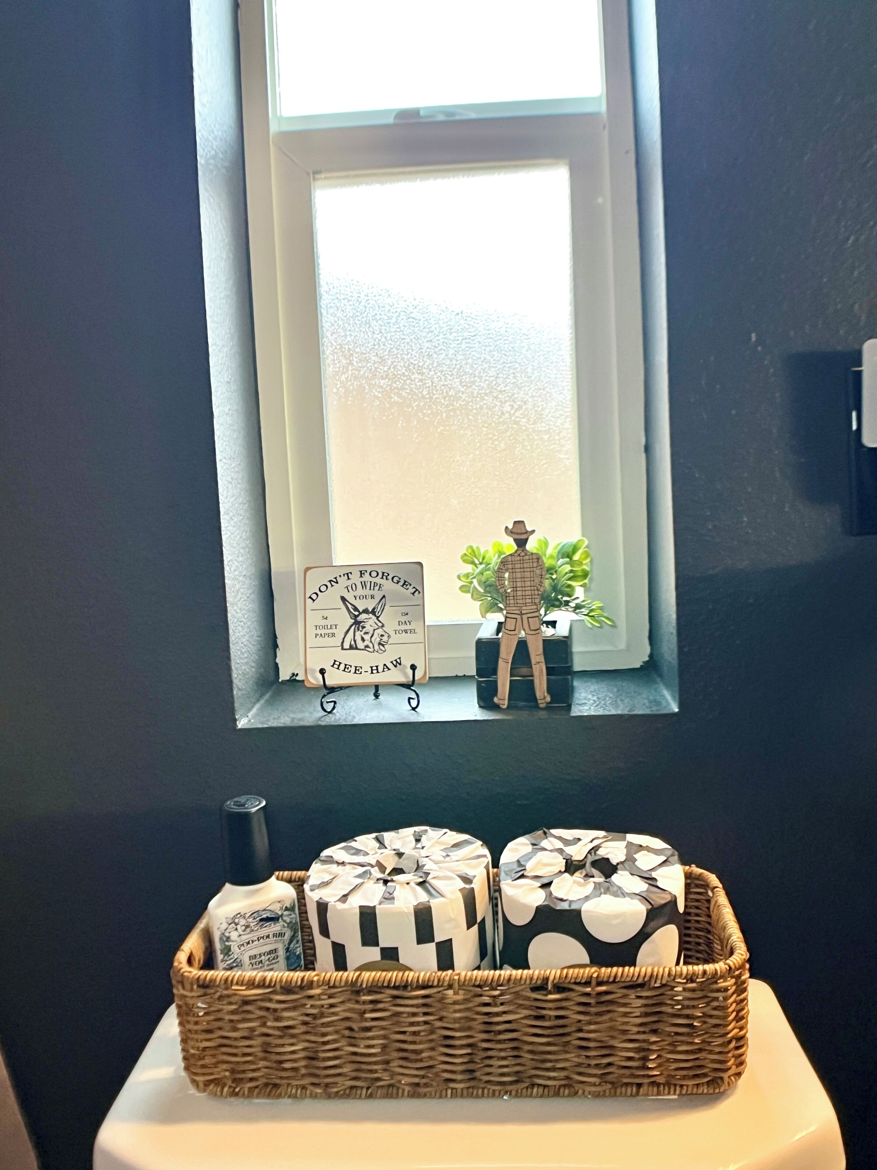 A bathroom with a window, a small plant, a decorative figure, and a basket of toiletries.